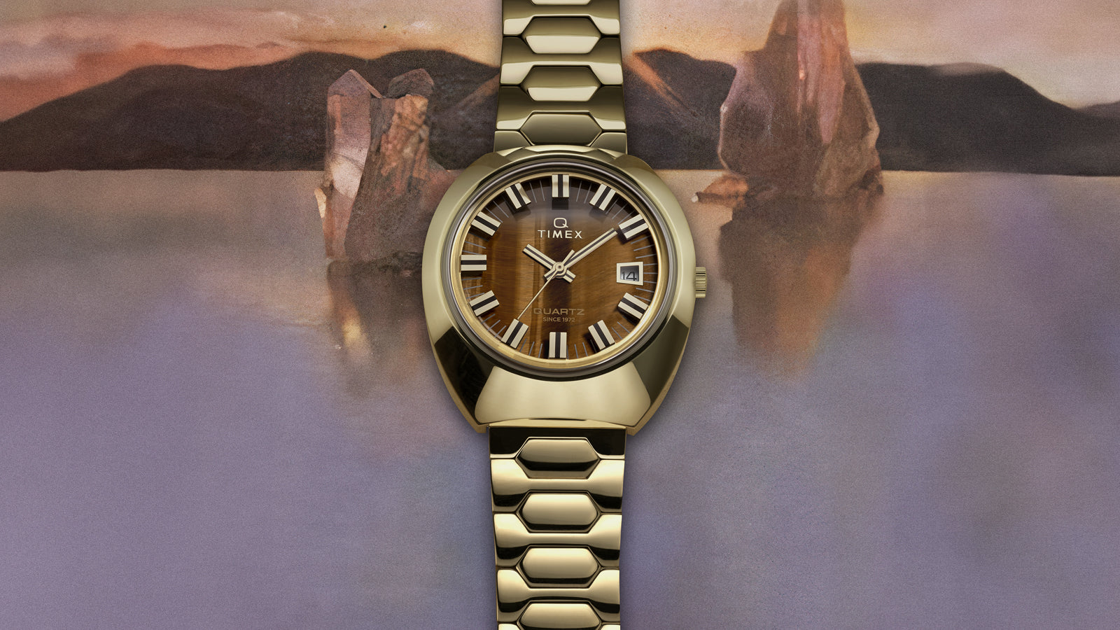 Q Timex® 1972 Time Machine Reissue wristwatch in polished gold-tone stainless steel with a faceted case and hexagonal-link bracelet. Features a tiger’s-eye style brown dial with silver-tone hour markers accented in black, luminous hands, and a framed date window at 3 o’clock, displayed against a textured, artistic background.