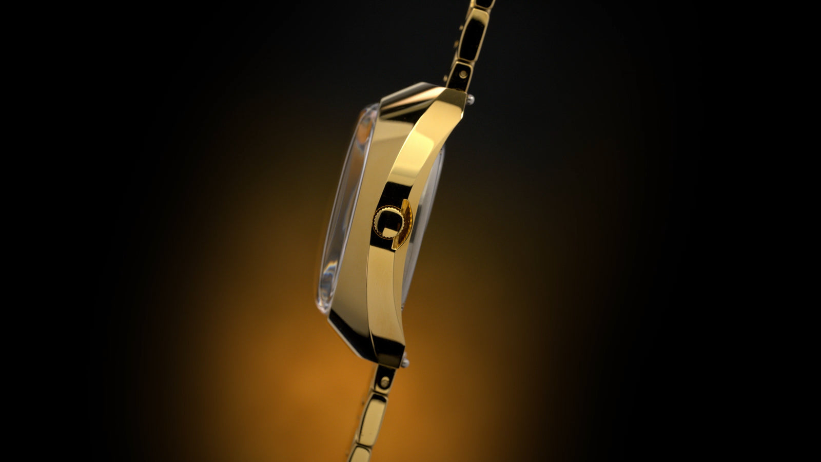 Side profile of the Q Timex® 1972 Time Machine Reissue gold-tone watch highlighting the faceted case, domed crystal, and gold-tone crown, with the hexagonal-link bracelet partially visible, set against a dark gradient background with warm amber lighting.