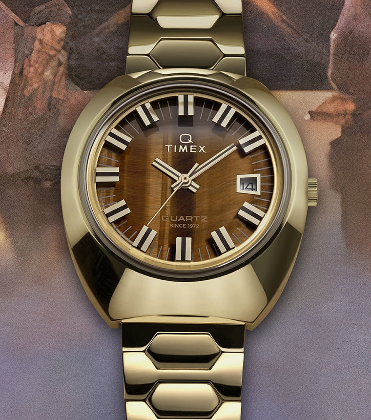 Q Timex® 1972 Time Machine Reissue wristwatch in polished gold-tone stainless steel with a faceted case and hexagonal-link bracelet. Features a tiger’s-eye style brown dial with silver-tone hour markers accented in black, luminous hands, and a framed date window at 3 o’clock, displayed against a textured, artistic background.