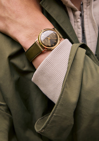 A close-up of a person wearing the Timex x Todd Snyder watch, featuring a green leather strap and gold-tone case, layered over a green jacket and beige hoodie in an outdoor urban setting.