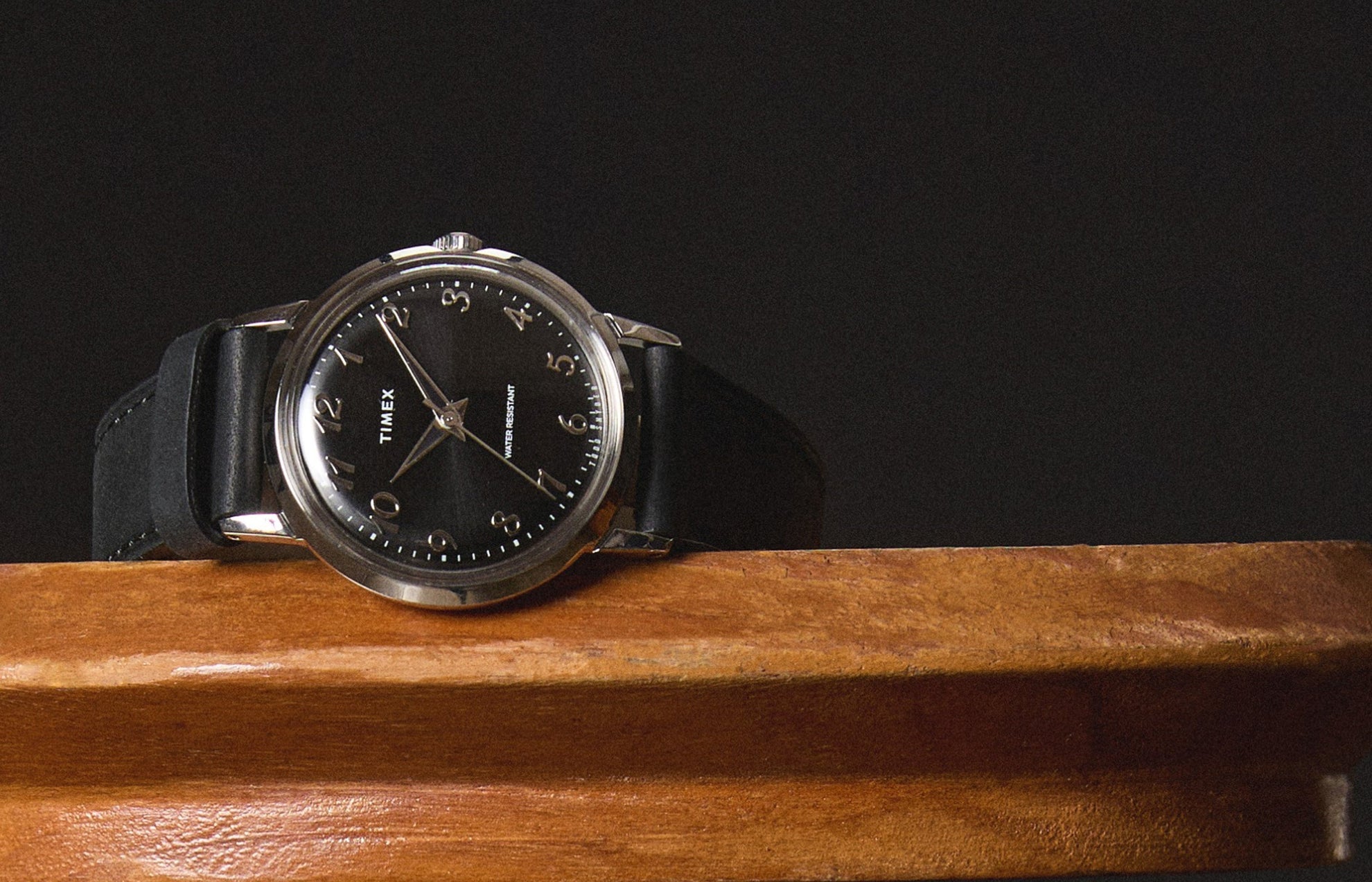 Timex x Todd Snyder watch resting on a wooden surface, highlighting the polished silver-tone case, black sunray dial, and classic Arabic numerals with a sleek black leather strap.