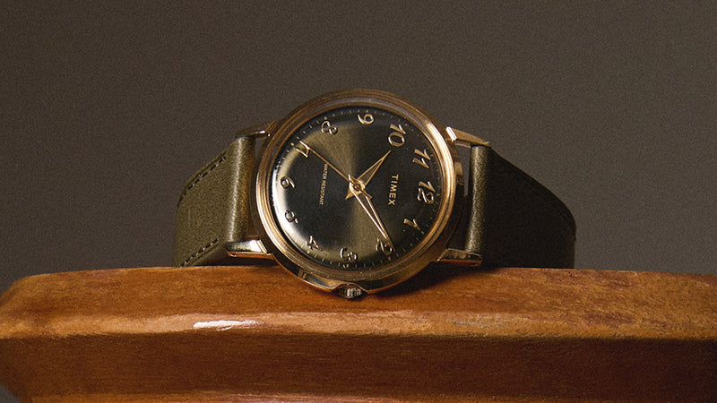 Timex x Todd Snyder watch resting on a wooden surface, showcasing the green dial, gold-tone case, and olive leather strap with a sophisticated, vintage-inspired look.