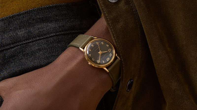 Lifestyle shot of a person wearing the Timex x Todd Snyder watch with a green strap, styled with a mustard sweater and brown suede jacket for a rich autumnal palette.