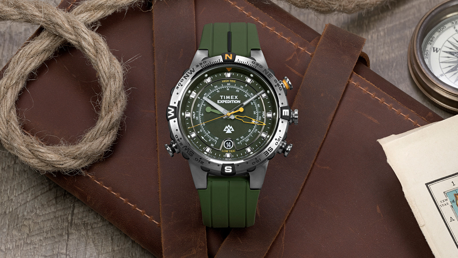 Timex Expedition® Tide-Temp-Compass watch with a green strap placed on a leather surface beside a vintage compass, nautical rope, and a folded map.