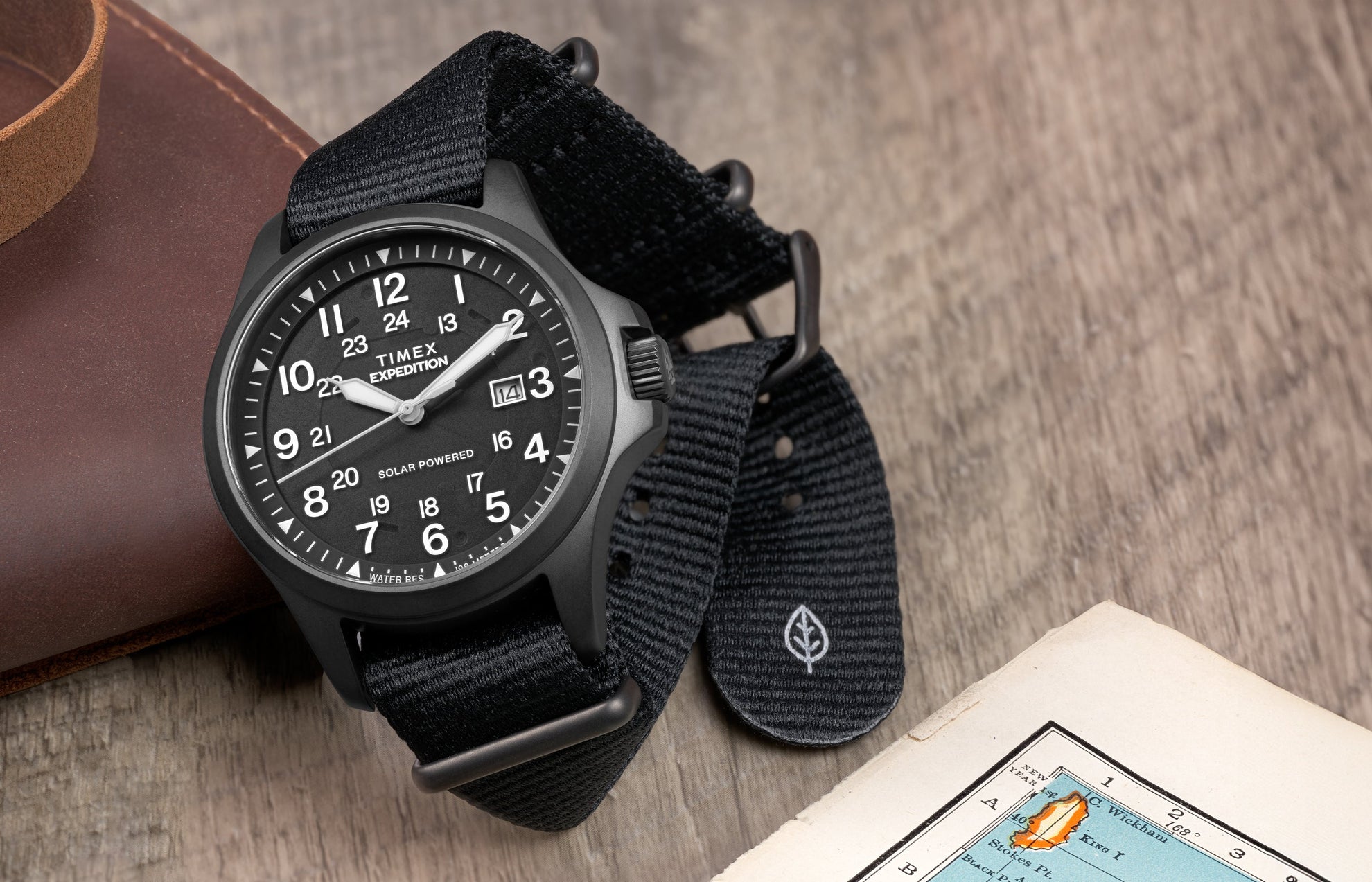Expedition Field Post Solar watch with black dial and strap on wood surface – Timex Expedition® Field Post Solar watch with matte black case, black dial, and black fabric strap, placed beside a leather pouch and vintage map.