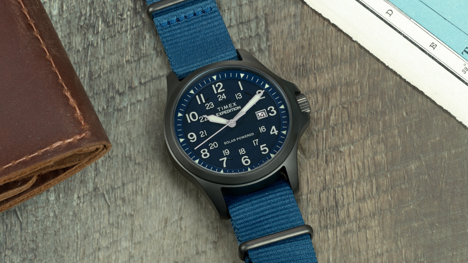 Expedition Field Post Solar watch with blue dial and strap on map background – Timex Expedition® Field Post Solar watch featuring a deep blue dial, white numerals, and fabric strap, placed on a wooden surface next to a vintage map.