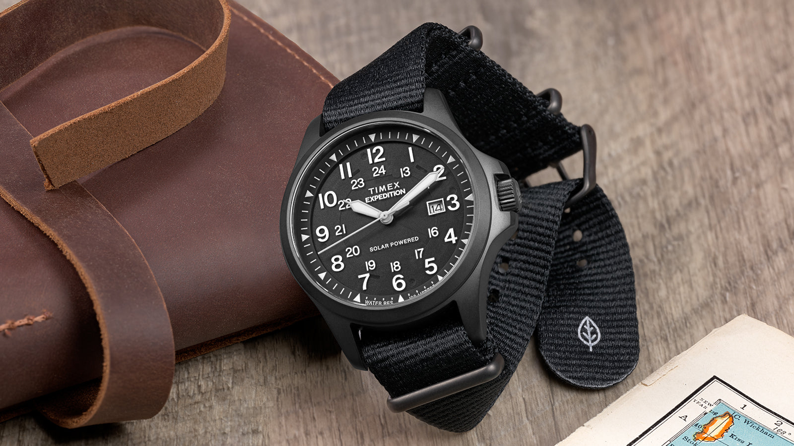 Expedition Field Post Solar watch with black dial and strap on wood surface – Timex Expedition® Field Post Solar watch with matte black case, black dial, and black fabric strap, placed beside a leather pouch and vintage map.