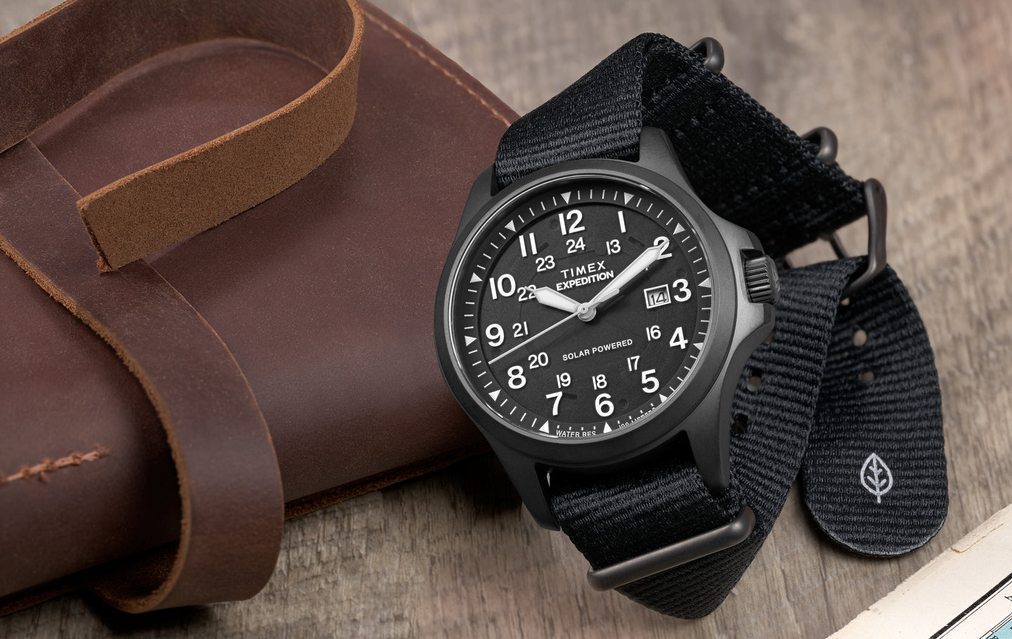 Timex Expedition® Field Post Solar black watch with recycled fabric strap, black dial, date display, and solar-powered movement on a leather journal.