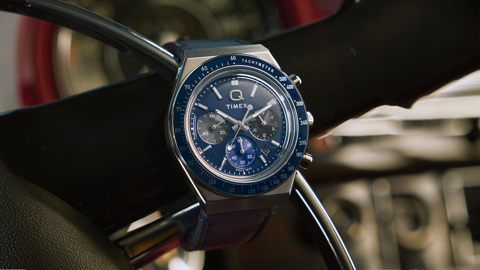 Q Timex® Chronograph on a steering wheel—blue dial, three sub-dials, tachymeter bezel, date at 4 o’clock.
