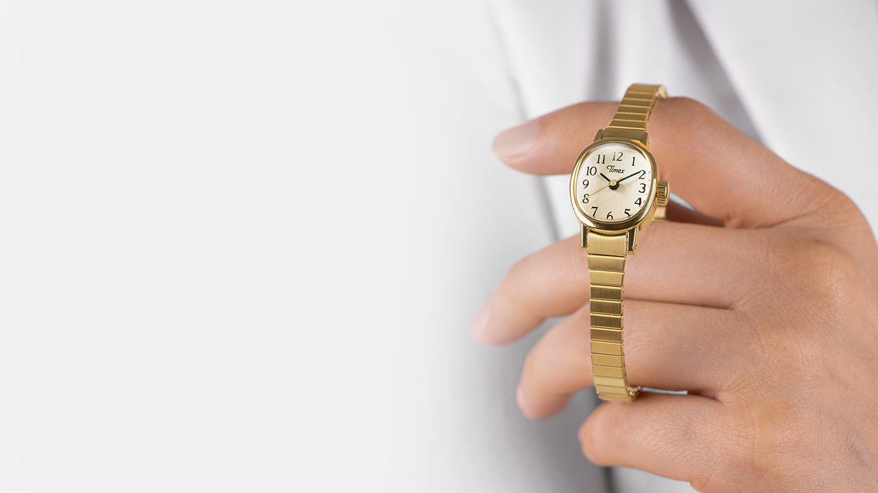 Close-up of a Timex Cavatina Mini gold-tone watch with white dial and black Arabic numerals, held delicately in hand.