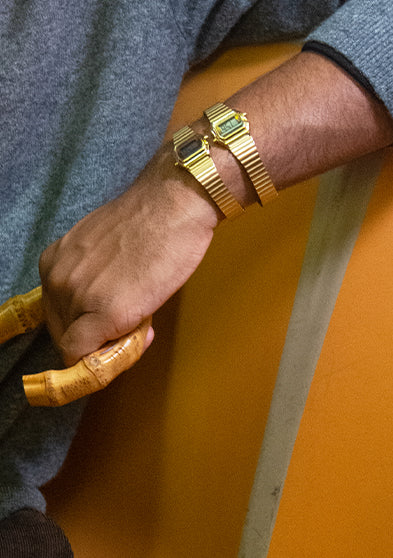 A person seated on a subway bench wearing two gold-tone Teeny Tiny Timex digital watches on one wrist. The watches complement layered gold necklaces and a bamboo-handled bag, reflecting Dimepiece’s nostalgic-meets-modern street style aesthetic.