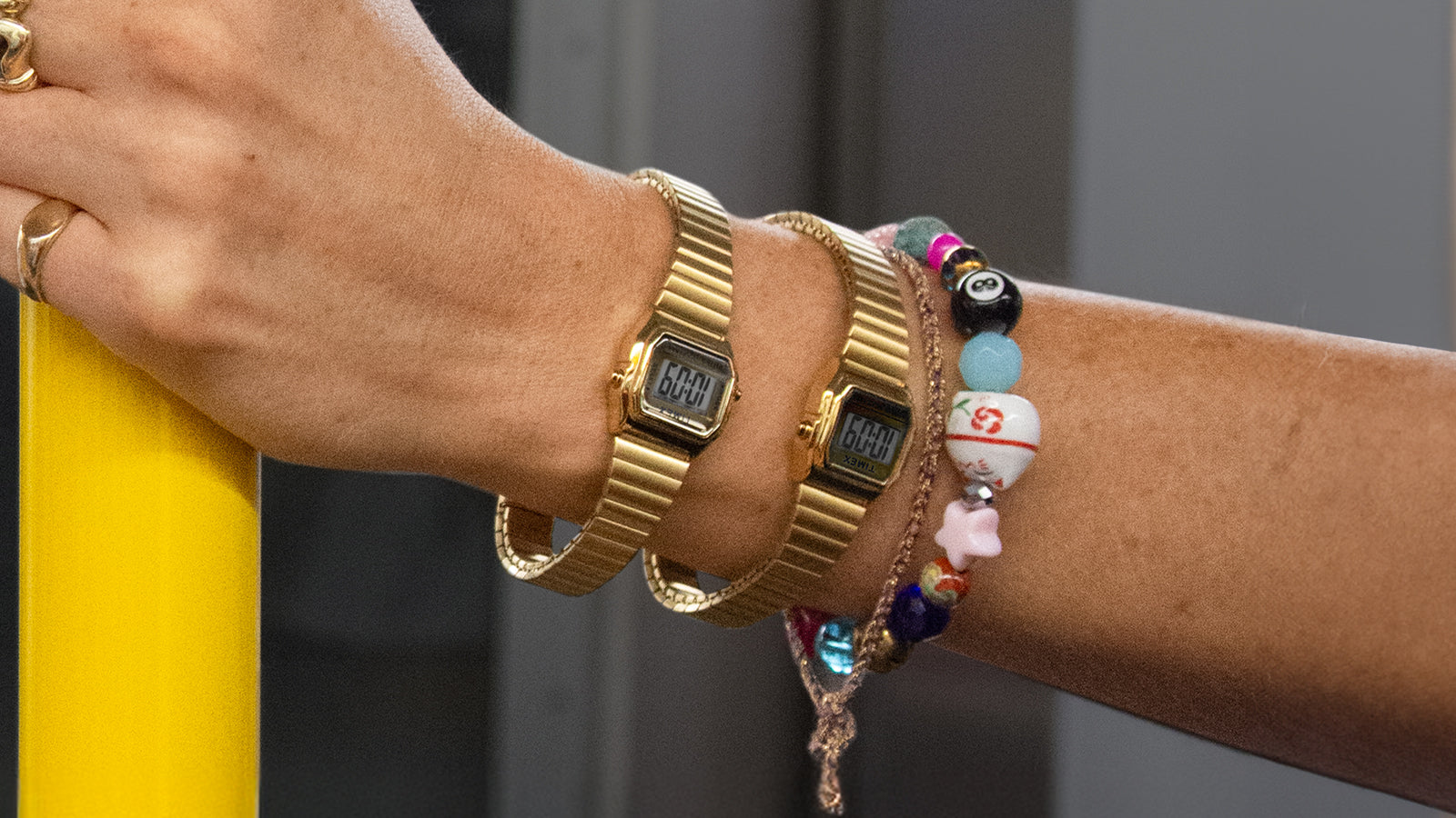 A person wearing two gold-tone Teeny Tiny Timex digital watches holds a yellow subway pole. The compact, retro-inspired watches are paired with colorful beaded bracelets and gold rings, capturing a playful and stylish city commute vibe.