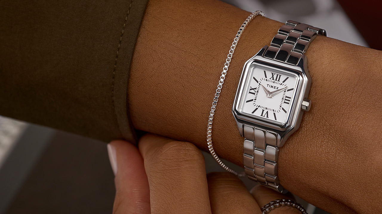 Timex Legacy Addison silver-tone square watch with white Roman-numeral dial, worn on the wrist beside a slim chain bracelet.