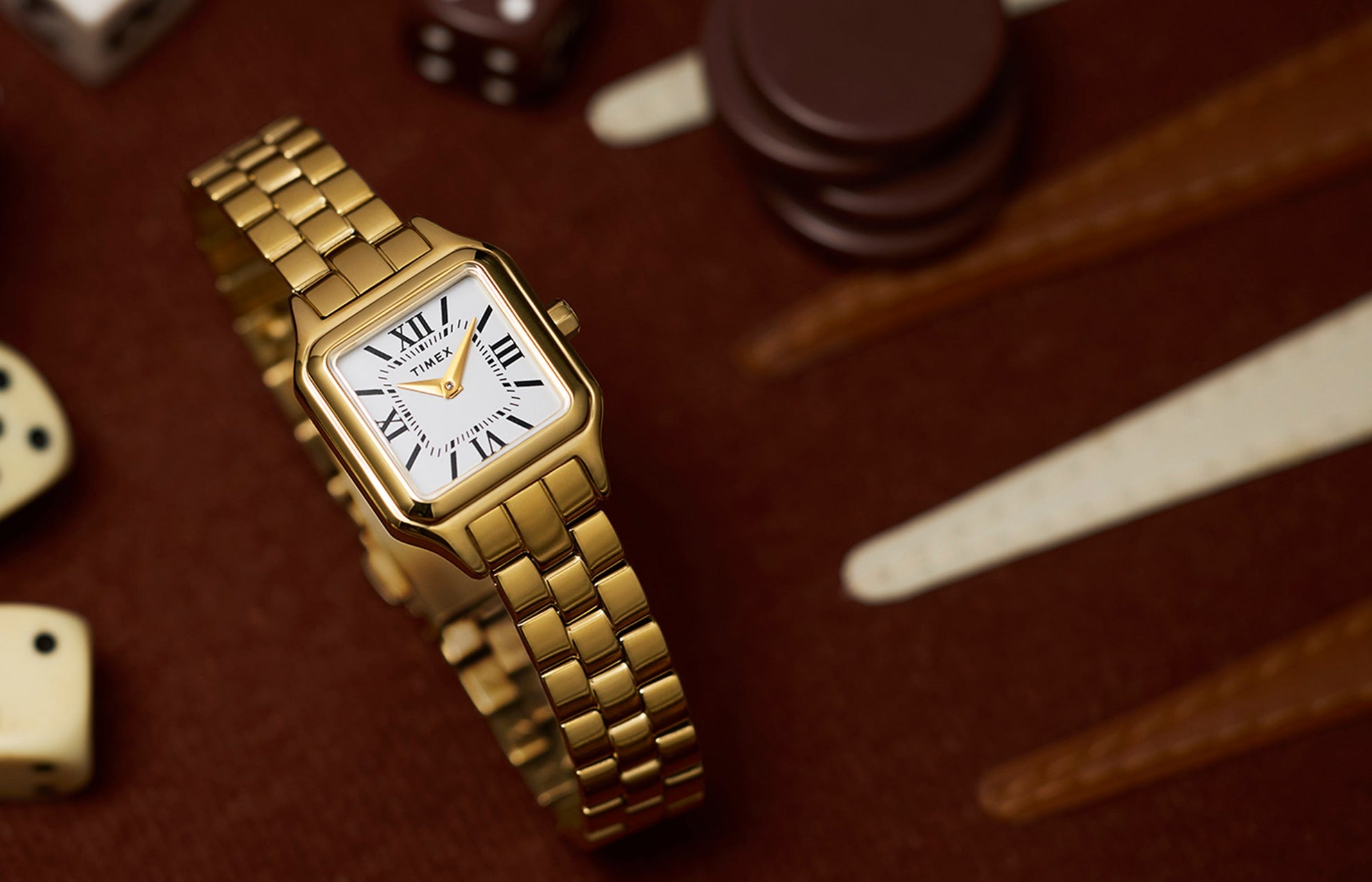 Timex Legacy Addison gold-tone women’s watch with rectangular case, white dial, and Roman numerals, styled on a backgammon board with dice.