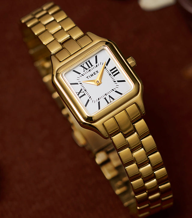 Close-up of the Timex Legacy Addison gold-tone watch showing square case, white dial with black Roman numerals, and gold hands on a link bracelet.