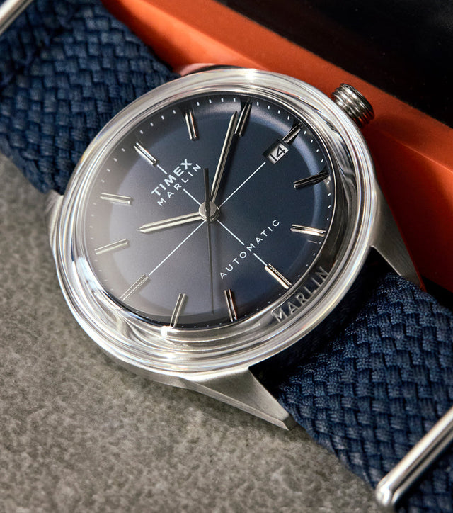 Timex Marlin® Jet Automatic watch with a gradient blue dial, stainless-steel case, and woven navy strap, displayed on a textured surface beside an orange and black object.