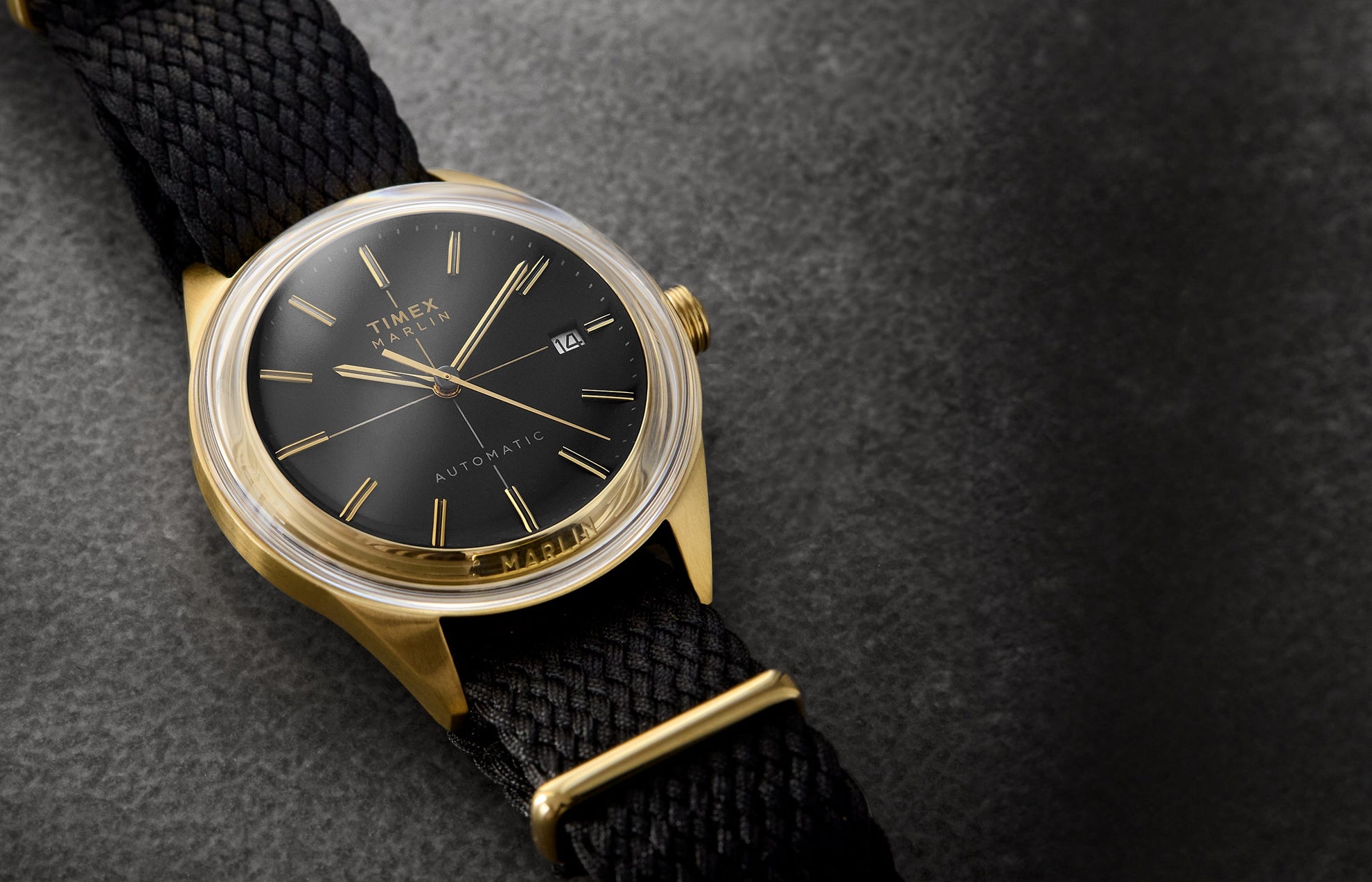 Timex Marlin® Jet Automatic watch with a black dial, gold-tone case, gold hands and indices, and black woven strap, displayed on a textured dark background.