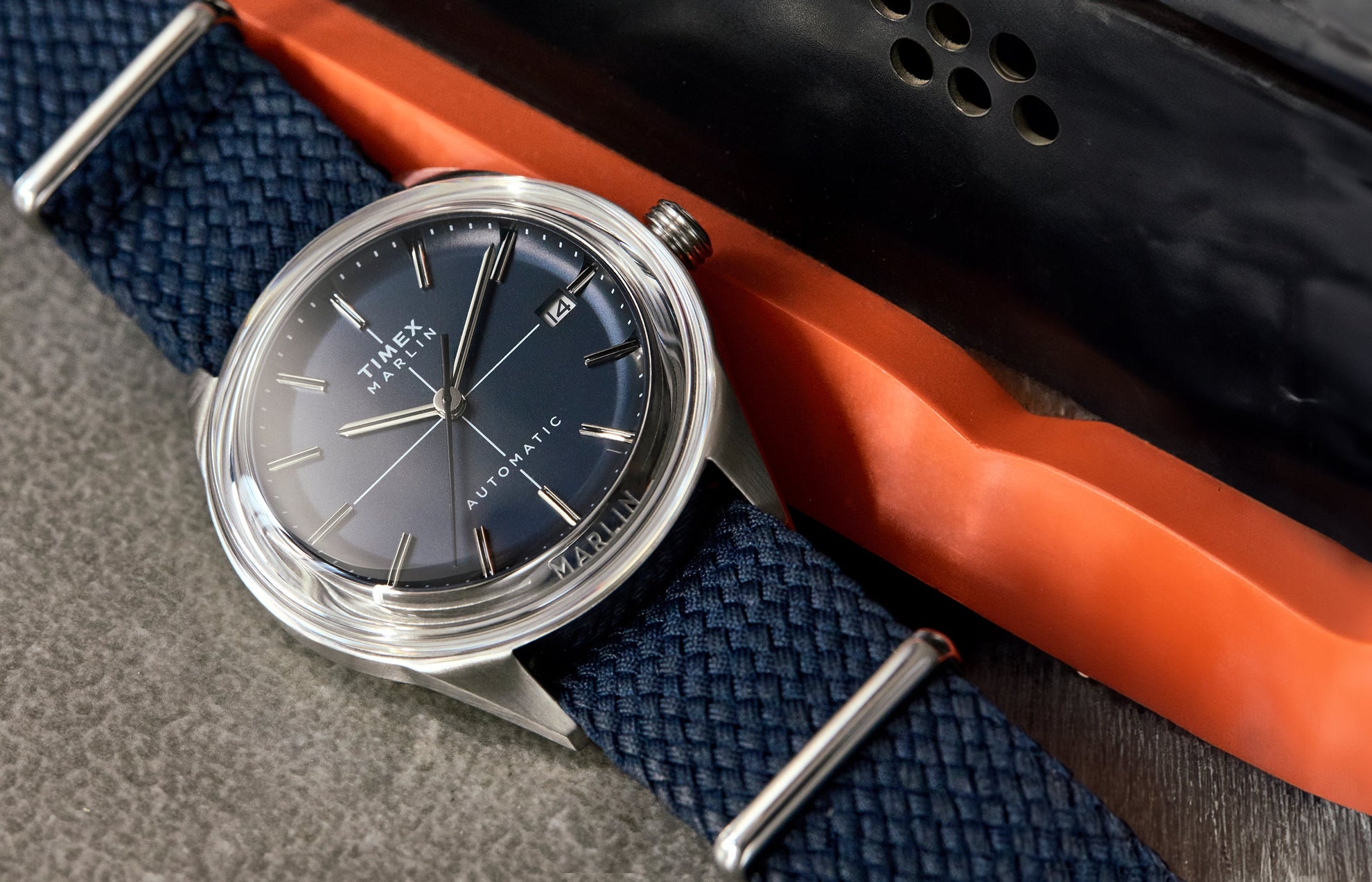 Timex Marlin® Jet Automatic watch with a gradient blue dial, stainless-steel case, and woven navy strap, displayed on a textured surface beside an orange and black object.