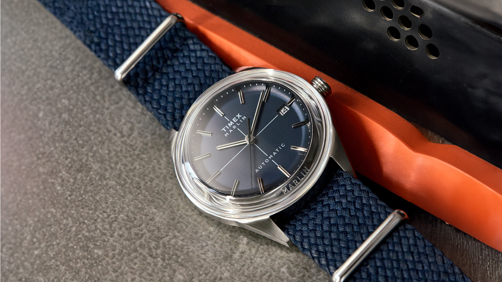 Timex Marlin® Jet Automatic watch with a gradient blue dial, stainless-steel case, and woven navy strap, displayed on a textured surface beside an orange and black object.