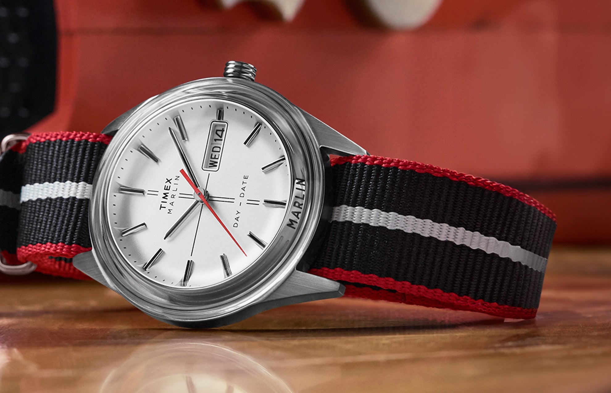 Timex Marlin® Jet Quartz watch with a silver dial, red seconds hand, and black-and-red striped fabric strap, displayed close-up on a wooden surface.