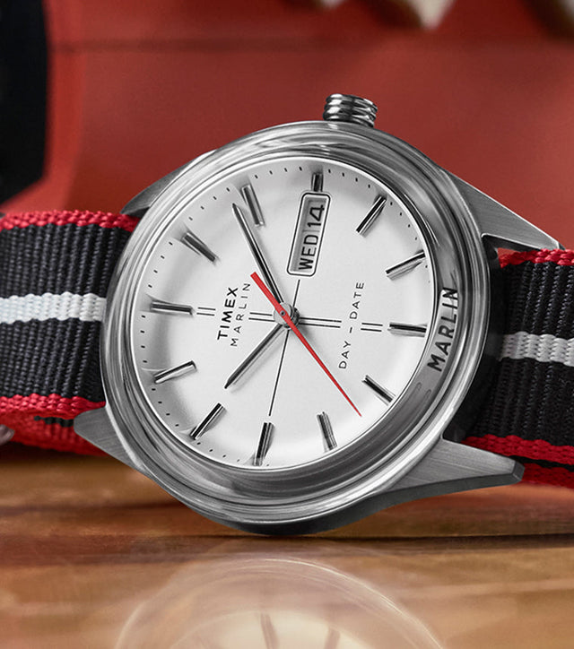 Timex Marlin® Jet Quartz watch with a silver dial, red seconds hand, and black-and-red striped fabric strap, displayed close-up on a wooden surface.