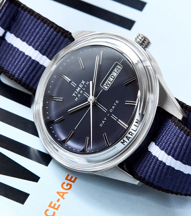 Timex Marlin® Jet Quartz watch with a deep blue dial, silver-tone markers, and day-date window, paired with a navy blue fabric strap featuring a white center stripe, displayed on a magazine cover.