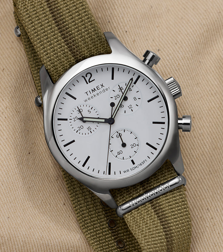 Timex Weekender New England Chronograph with white dial and three sub-dials on an olive fabric strap, styled on tan canvas.