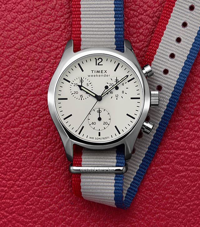 Timex Weekender New England Chronograph watch with a white dial, black hands, and a red, white, and blue strap, displayed on a red textured background.