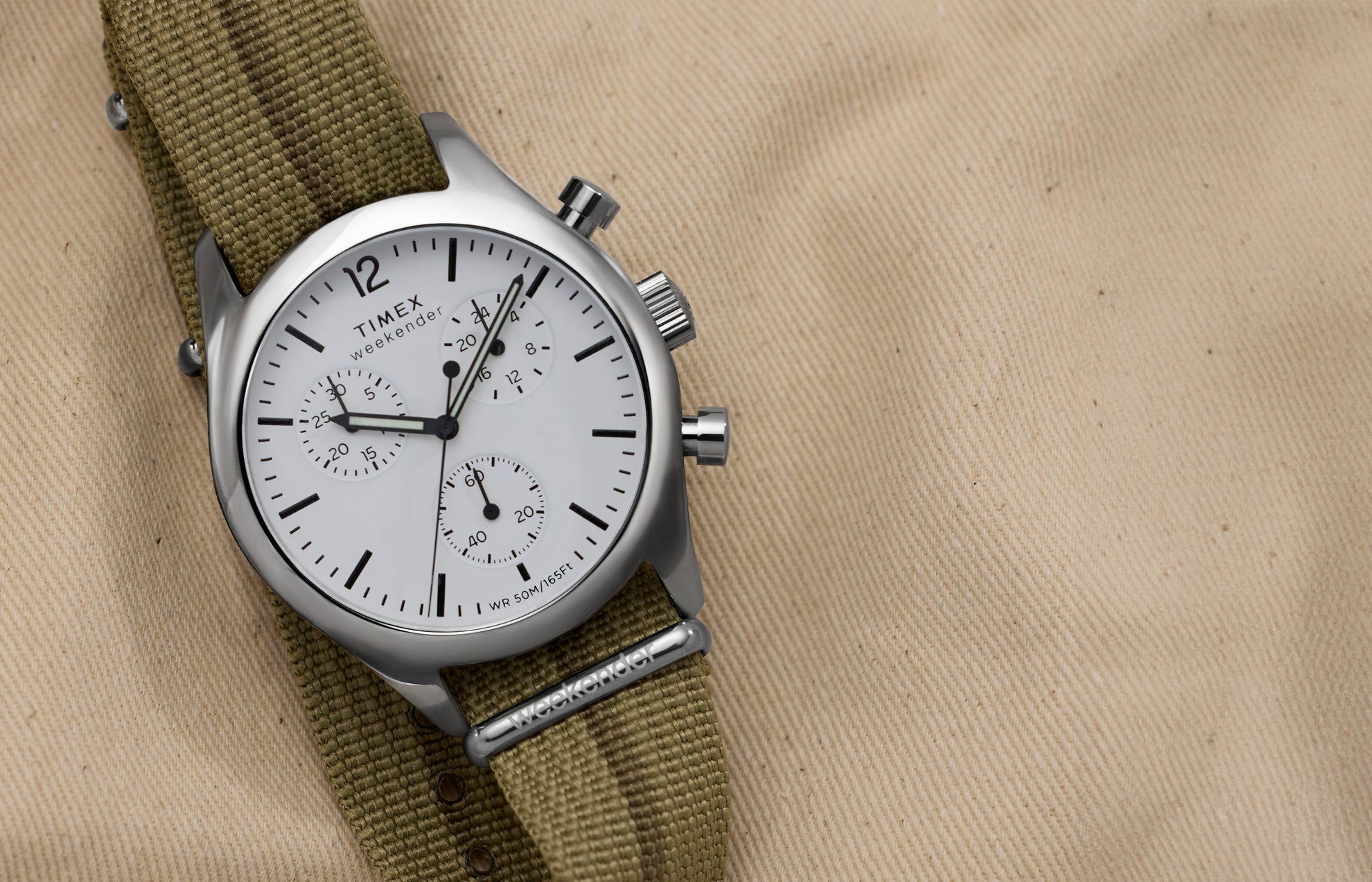 Timex Weekender New England Chronograph with white dial and three sub-dials on an olive fabric strap, styled on tan canvas.