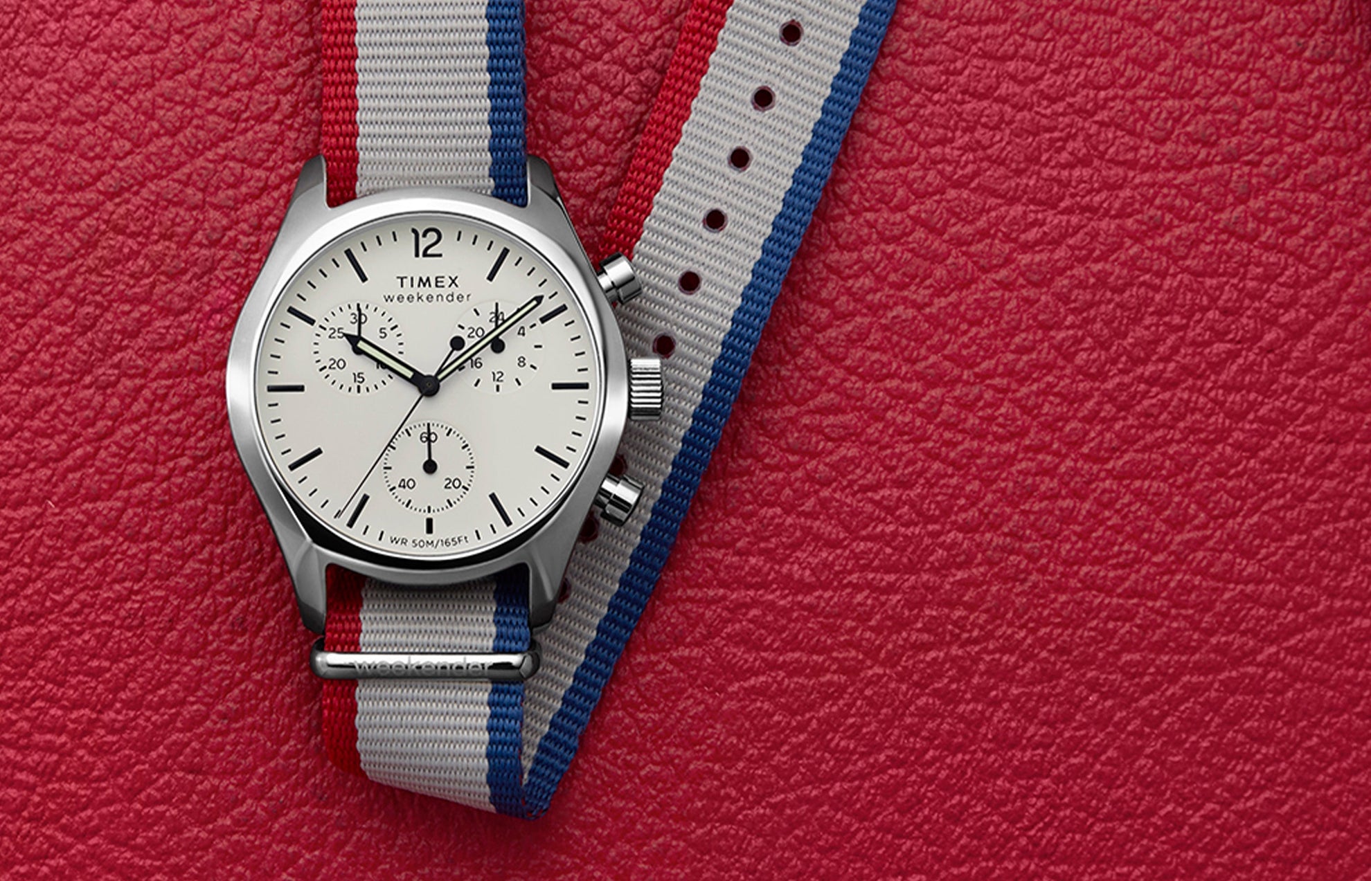 Timex Weekender New England Chronograph watch with a white dial, black hands, and a red, white, and blue strap, displayed on a red textured background.