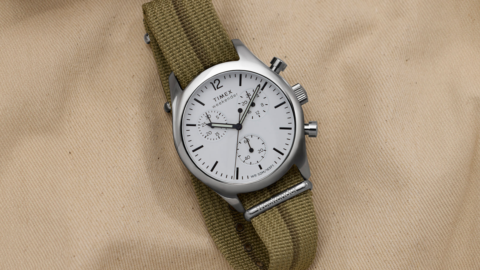 Timex Weekender New England Chronograph with white dial and three sub-dials on an olive fabric strap, styled on tan canvas.