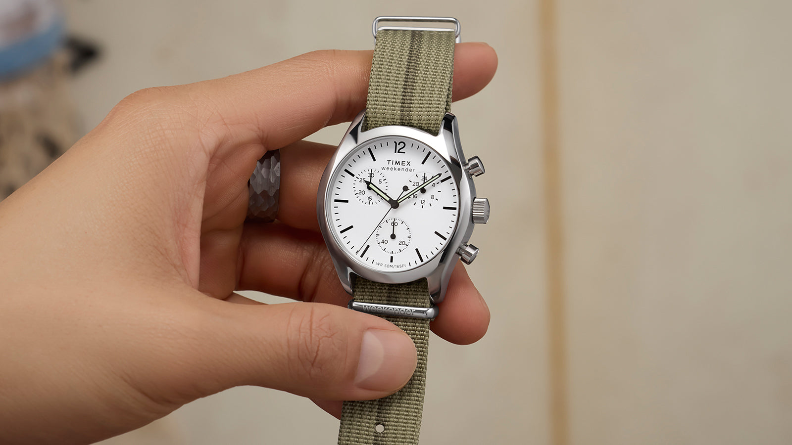 Hand holding a Timex Weekender New England Chronograph with white dial, three sub-dials, stainless-steel case, and olive fabric strap.