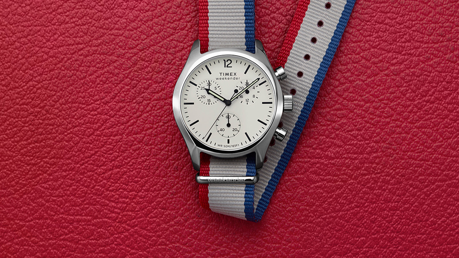 Timex Weekender New England Chronograph watch with a white dial, black hands, and a red, white, and blue strap, displayed on a red textured background.
