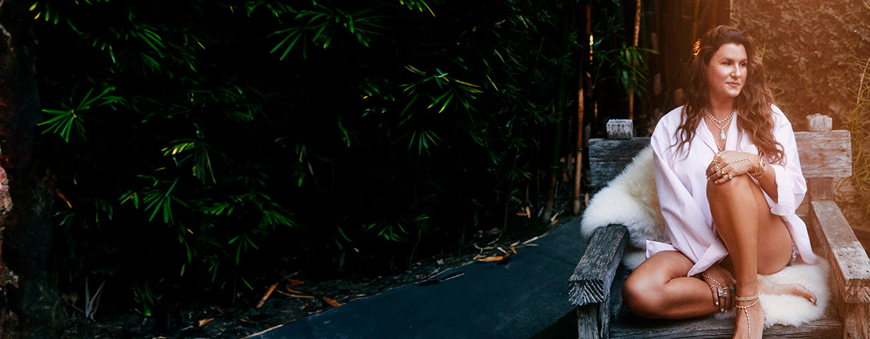 Woman seated on a rustic wooden bench with a white fur throw, wearing layered jewelry and a light robe-style outfit, surrounded by lush greenery in a relaxed outdoor setting.