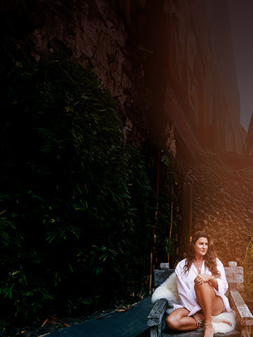 Woman seated on a rustic wooden bench with a white fur throw, wearing layered jewelry and a light robe-style outfit, surrounded by lush greenery in a relaxed outdoor setting.