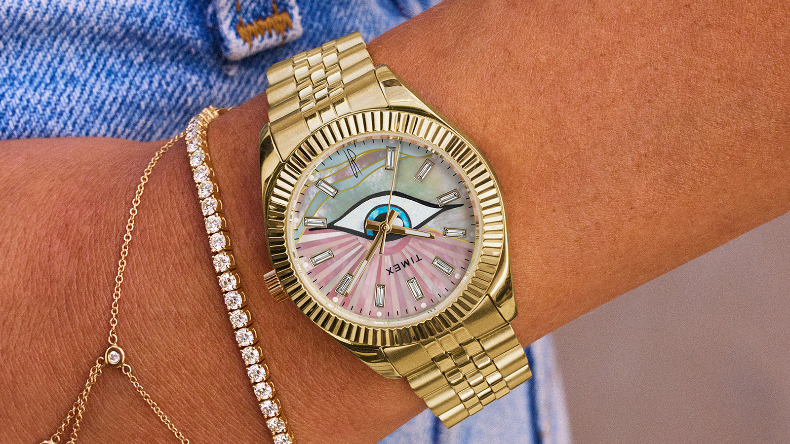 Close-up of a wrist wearing the Timex x Jacquie Aiche gold watch with a multicolored mother-of-pearl dial featuring an eye motif, styled alongside delicate gold and diamond bracelets, against a denim background.