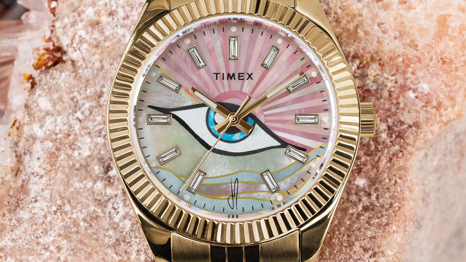 Detailed close-up of the Timex x Jacquie Aiche watch dial, showcasing the pink and iridescent mother-of-pearl face with an illustrated eye motif, gold-tone hands, and baguette-style crystal hour markers in a gold case.