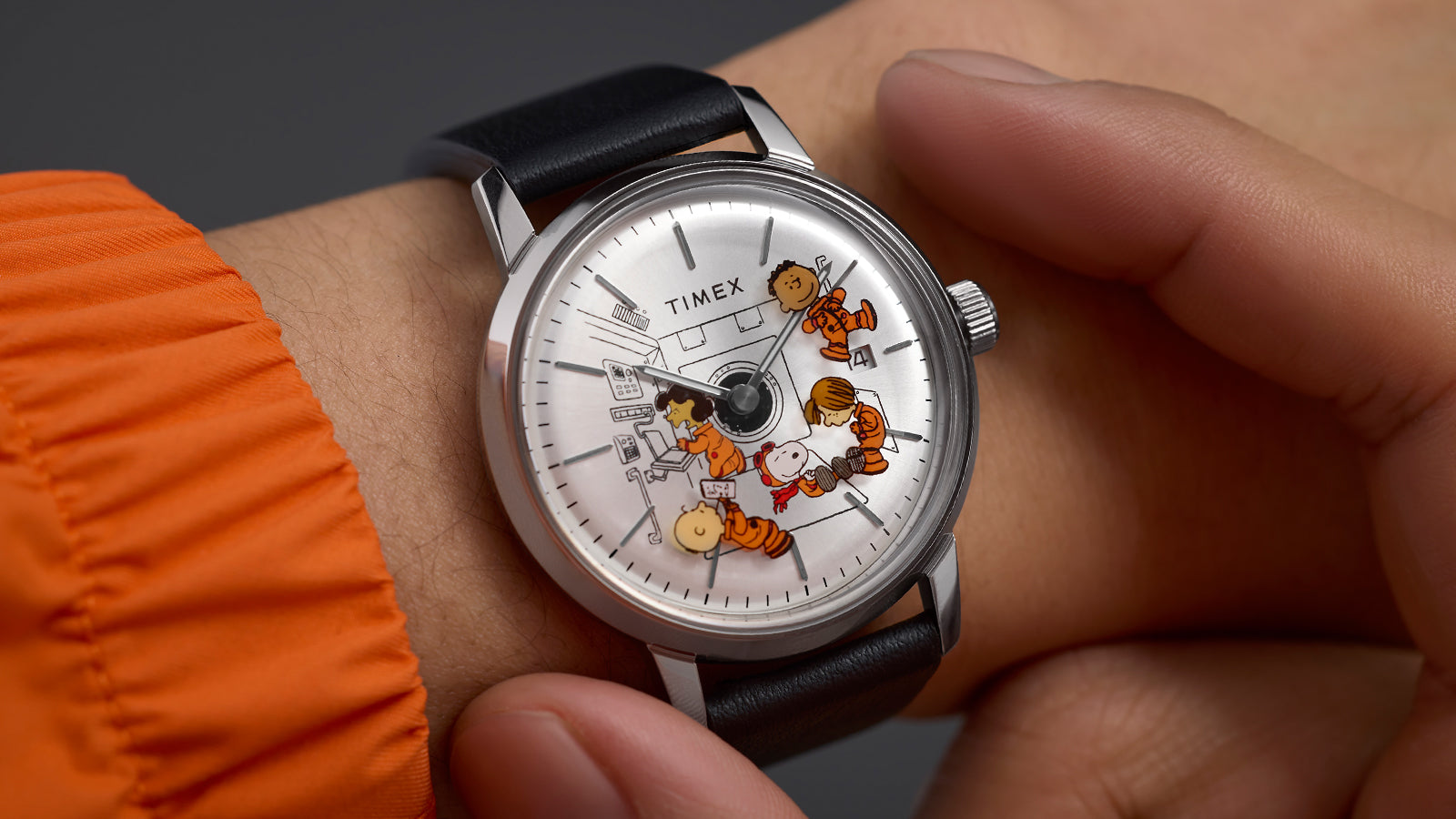 Close-up of the Timex x Peanuts® Marlin® Automatic watch worn on a wrist. The silver dial displays Peanuts characters in orange space suits floating in zero gravity, with black hour markers and a date window at 3 o’clock.