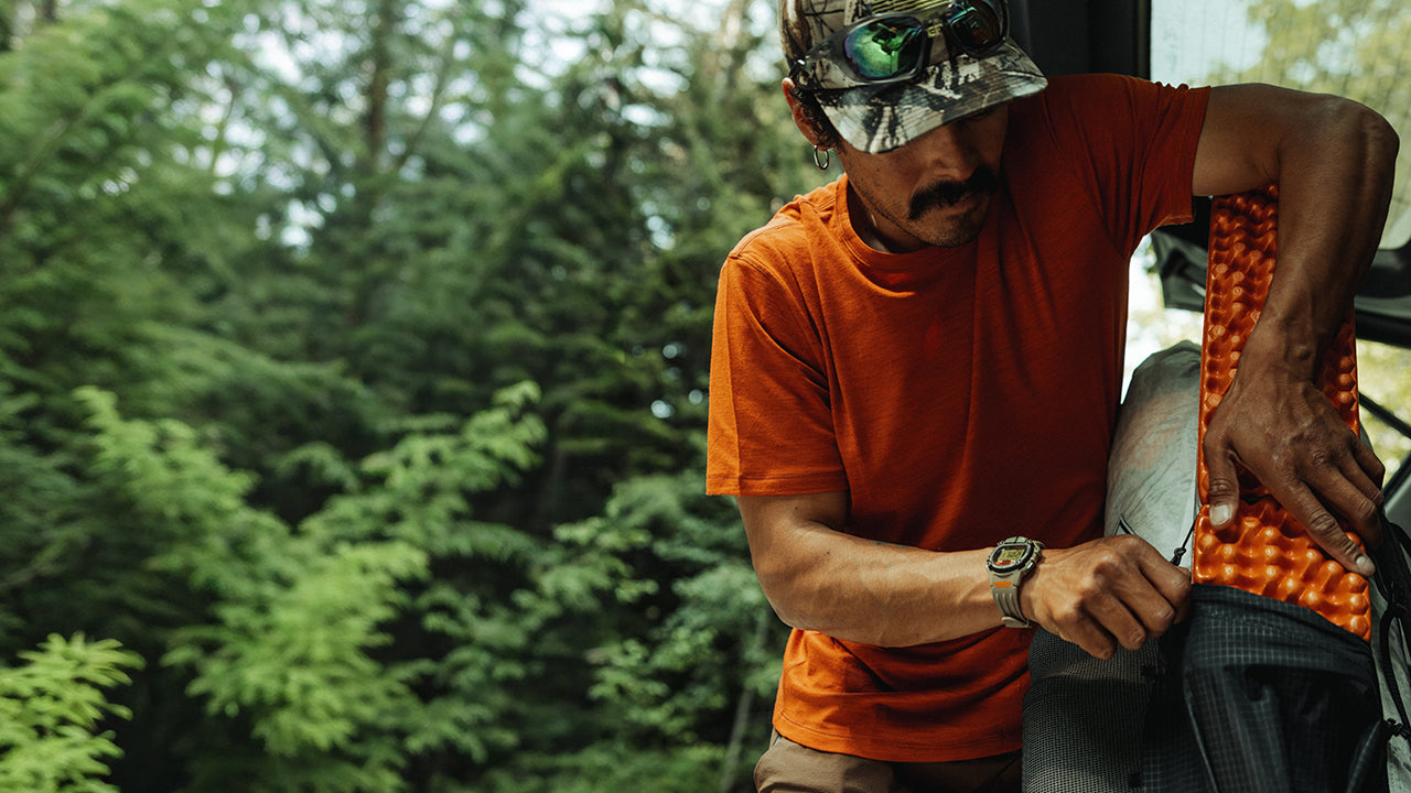A man in an orange shirt and cap wears the TIMEX® IRONMAN® Flix x Huckberry while outdoors, packing camping gear. The rugged watch is shown as part of his active outdoor lifestyle.
