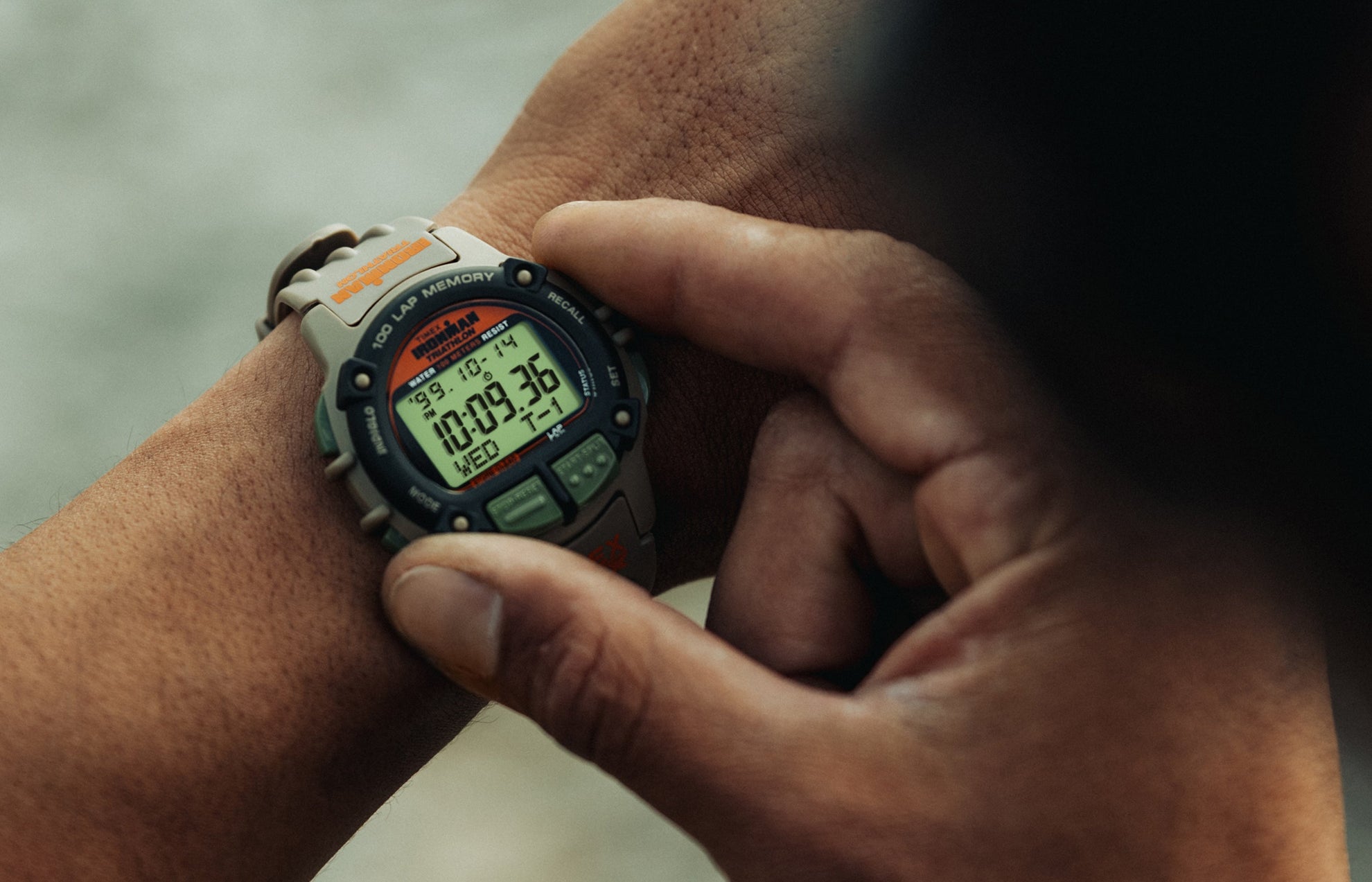 A close-up of a person wearing the TIMEX® IRONMAN® Flix x Huckberry digital sports watch, showing the rugged khaki case, black bezel, and green function buttons. The digital display reads 10:09:36 with the date Wednesday 10-14.