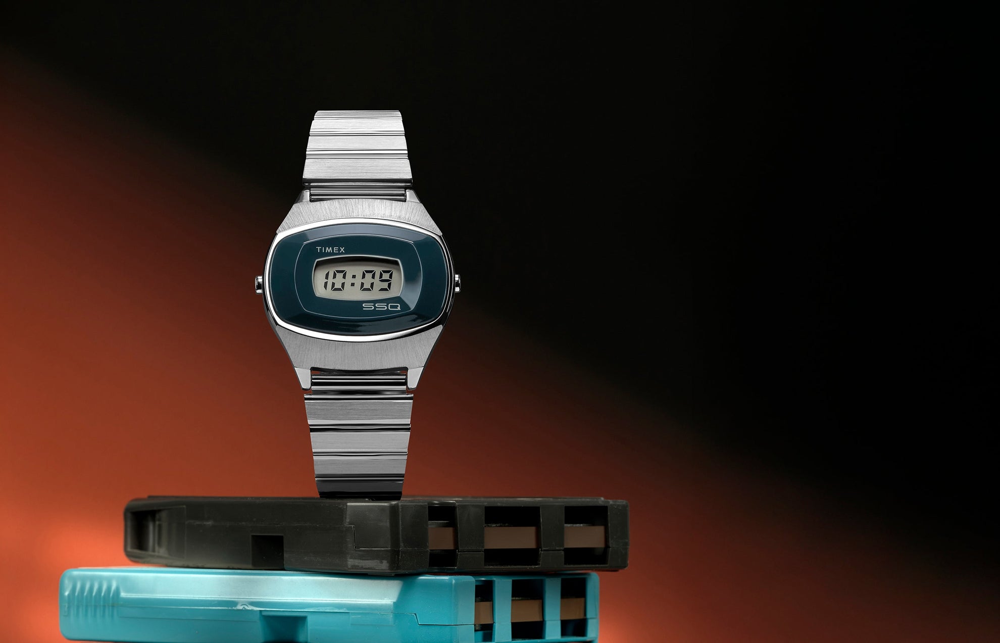 Q Timex® 1975 SSQ Digital Reissue watch displayed upright on stacked retro cartridges against a black and orange gradient background, showcasing its stainless-steel case and blue digital dial.