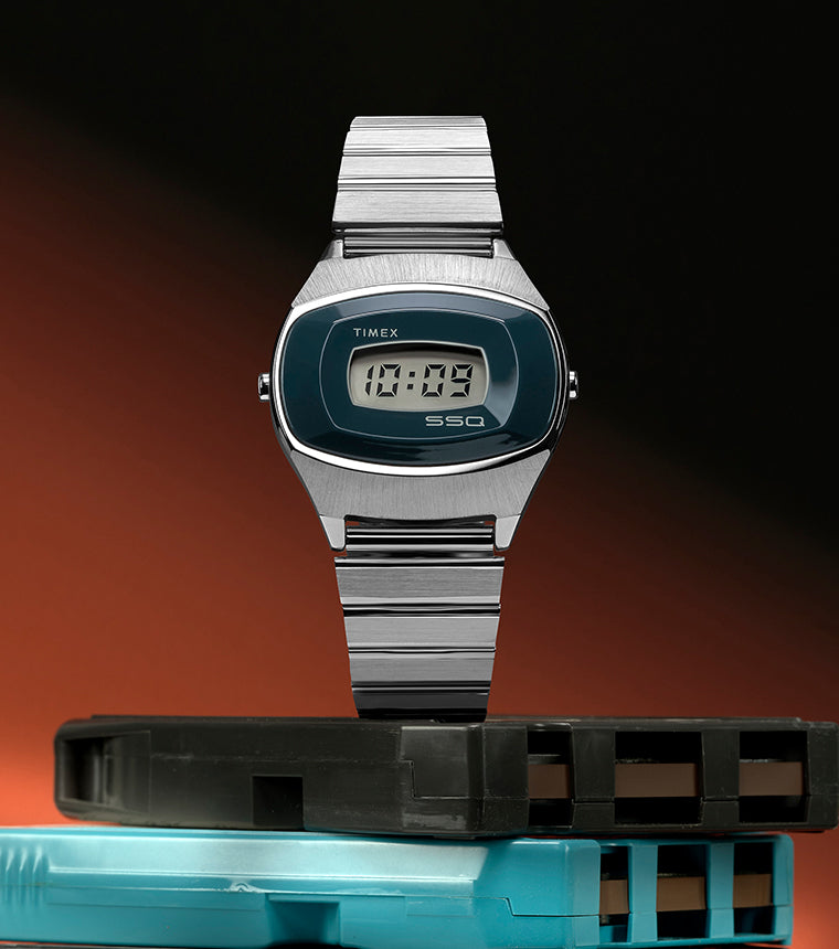 Q Timex® 1975 SSQ Digital Reissue watch displayed upright on stacked retro cartridges against a black and orange gradient background, showcasing its stainless-steel case and blue digital dial.
