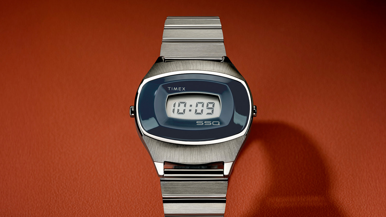 Centered product shot of the Q Timex® 1975 SSQ Digital Reissue on an orange background emphasizing its brushed steel finish, sleek integrated bracelet, and retro digital style.