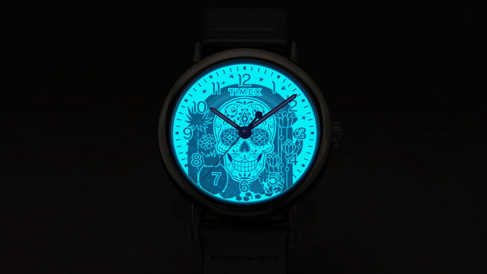 Timex Weekender Day of the Dead watch dial glowing in the dark with INDIGLO® backlight, revealing a bright blue sugar skull and floral details.