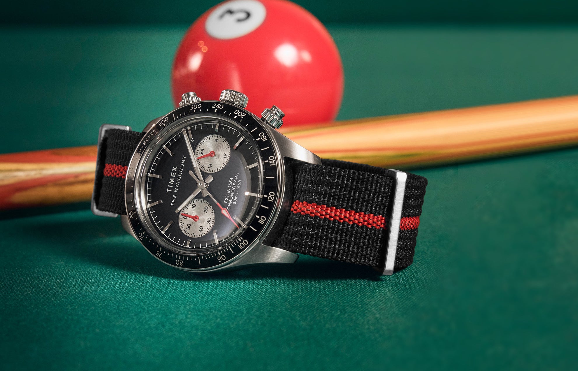 Timex Waterbury Heritage Chronograph watch with a black dial, silver subdials, and a black fabric strap with a red stripe, placed on a pool table beside a cue stick and billiard ball.