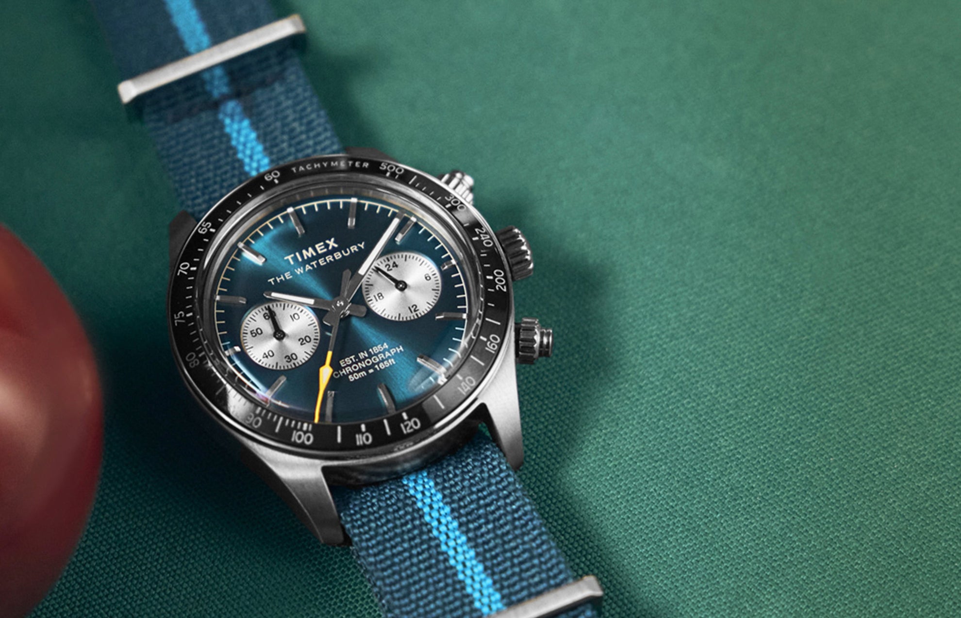 Timex Waterbury Heritage Chronograph with a blue sunray dial, silver subdials, and blue fabric strap featuring a light blue stripe, displayed on a pool table.