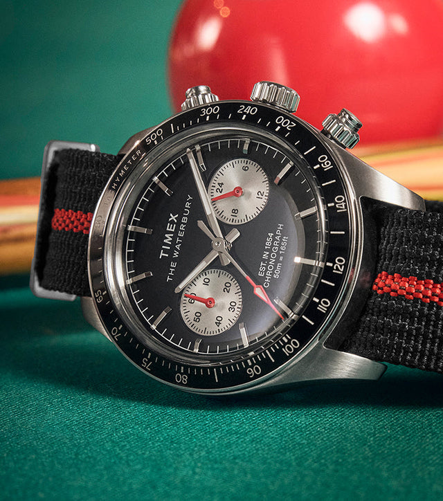 Timex Waterbury Heritage Chronograph watch with a black dial, silver subdials, and a black fabric strap with a red stripe, placed on a pool table beside a cue stick and billiard ball.