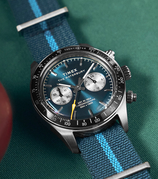 Timex Waterbury Heritage Chronograph with a blue sunray dial, silver subdials, and blue fabric strap featuring a light blue stripe, displayed on a pool table.