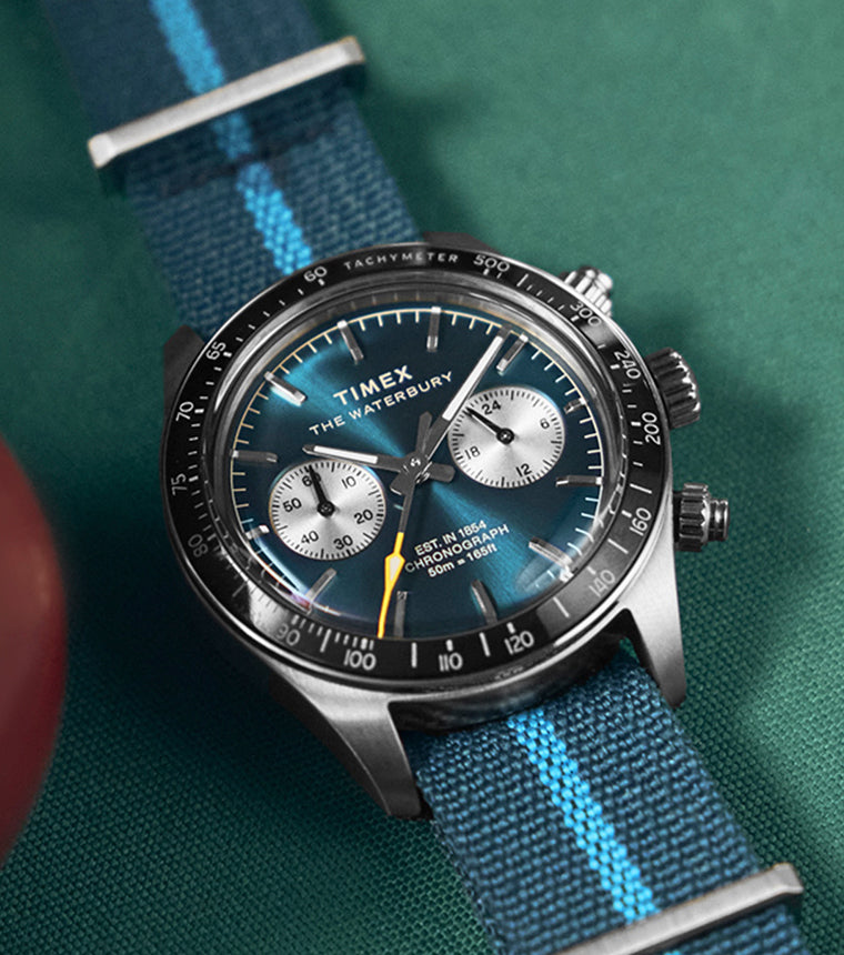 Timex Waterbury Heritage Chronograph with a blue sunray dial, silver subdials, and blue fabric strap featuring a light blue stripe, displayed on a pool table.