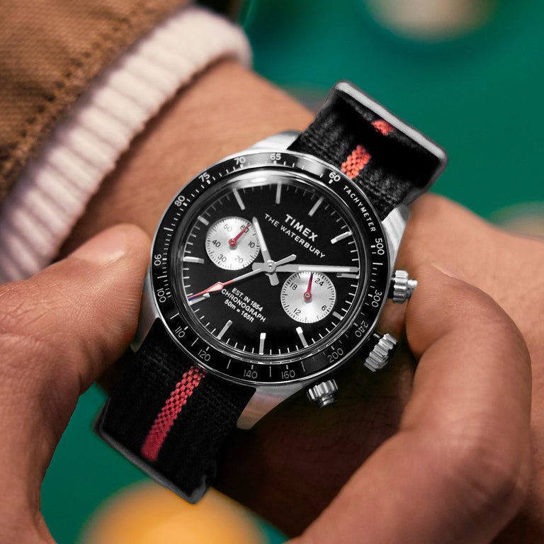 Timex Waterbury Heritage Chronograph worn on wrist, showing its black dial with silver subdials and red accents, secured by a black and red fabric strap.